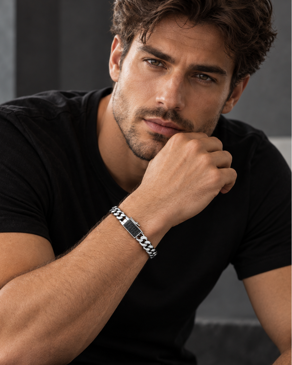 A man wearing a black t-shirt and black pants, resting his chin on his hand, showcasing a sterling silver bracelet with a black diamond pavé clasp on his wrist.